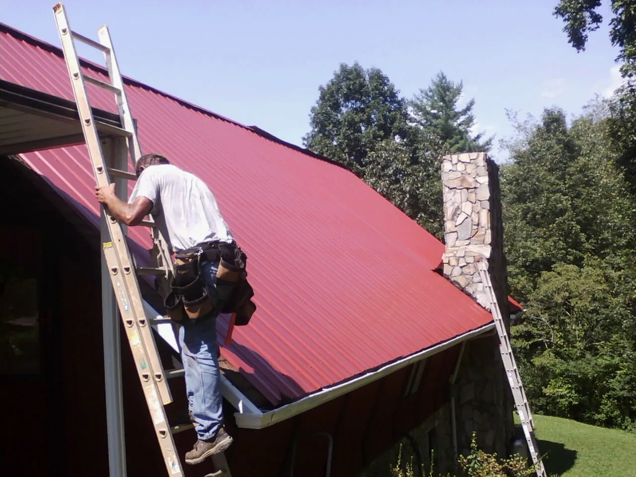 Roofer on a ladder beside a red metal roof and stone chimney — Soffit & Fascia Repair by Roofing Brothers in Coal Center